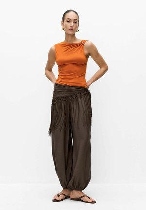 Femme debout, mains dans le dos, portant un haut orange sans manches, un pantalon large marron avec des détails en franges et des sandales marron sur fond blanc.
