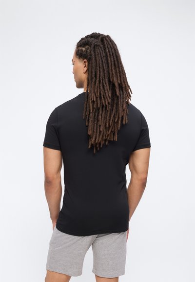 Άνδρας με μακριά ντραντλοκς που φοράει μαύρο μπλουζάκι και γκρι σορτς, στέκεται με την πλάτη προς την κάμερα σε απλό φόντο.