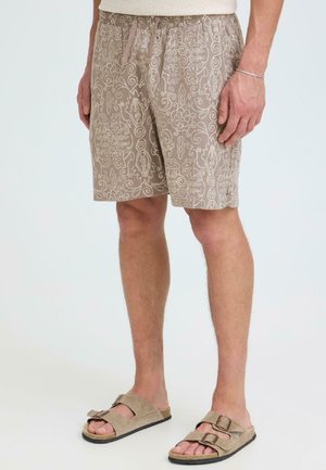 Homme portant un short beige à motifs floraux avec taille à cordon et sandales beige à double bride sur un fond uni.