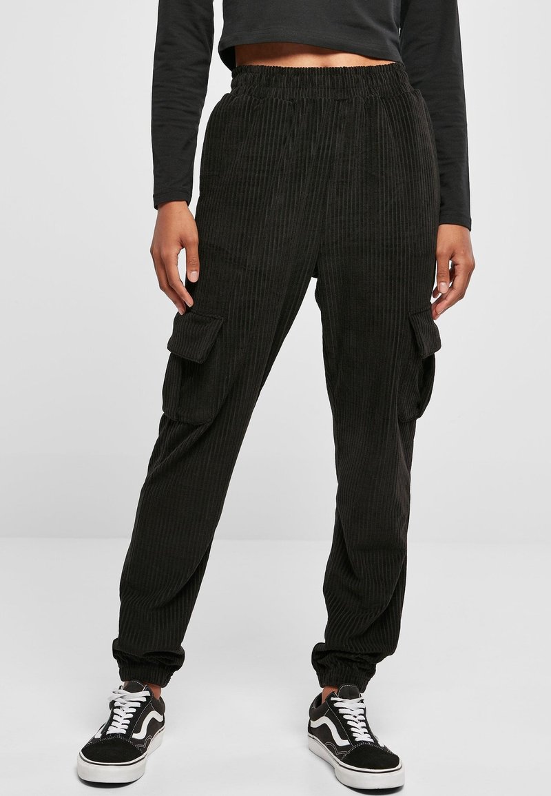 Femme portant un pantalon cargo côtelé noir avec taille et poignets élastiques, associé à des baskets noires et blanches sur un fond uni.