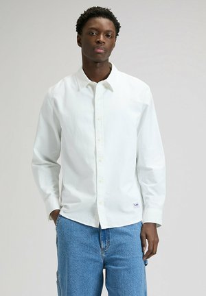 Młody mężczyzna z krótkimi kręconymi włosami, ubrany w białą koszulę z długim rękawem zapinaną na guziki oraz niebieskie dżinsy, stojący na jednolitym tle.