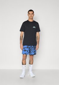 Sort t-shirt med korte ærmer og grønt logo, parret med blåmønstrede shorts, hvide sokker og hvide sportssko.