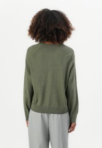 Maglione verde oliva con una finitura testurizzata, maniche raglan e fondo a coste. Indossato sopra pantaloni grigi chiari a righe.