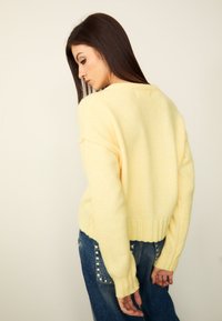 Pull léger jaune tricoté avec une coupe décontractée, ourlet et poignets côtelés, porté avec un jean denim bleu foncé orné de détails cloutés.