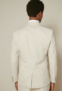 Veste de costume beige clair, tissu texturé, fente simple à l'arrière, boutons assortis et revers traditionnels.