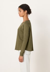 Kihara juustega naine, kes kannab oliivrohelist pikade varrukatega särki ja helesiniseid teksasid, seisab külgvaates tavalise tausta ees.