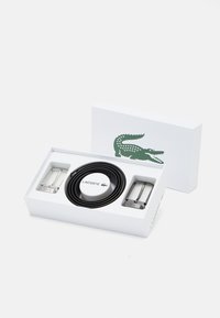 Cintura in pelle nera, fibbia circolare bianca con logo, fibbie in metallo argentato, presentata in una scatola bianca con logo di coccodrillo verde.