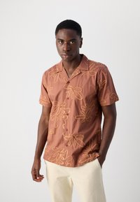 Camicia a maniche corte in tessuto ruggine con ricami floreali gialli, dotata di colletto, chiusura con bottoni e vestibilità rilassata. Tessuto morbido.