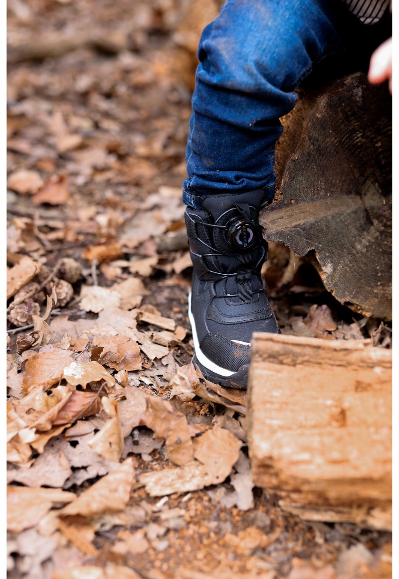 Schwarzer Schneestiefel mit strukturiertem Obermaterial, integriertem Schnürsystem und weißer Sohle, vor einem Hintergrund aus Blättern und Holzstämmen.