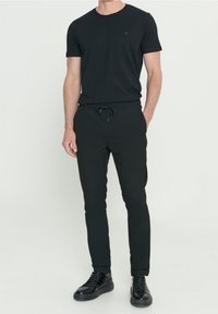 Man in een zwart T-shirt met korte mouwen, zwarte broek met tuniekkoord en zwarte sneakers, staand met één hand in zijn zak tegen een witte achtergrond.