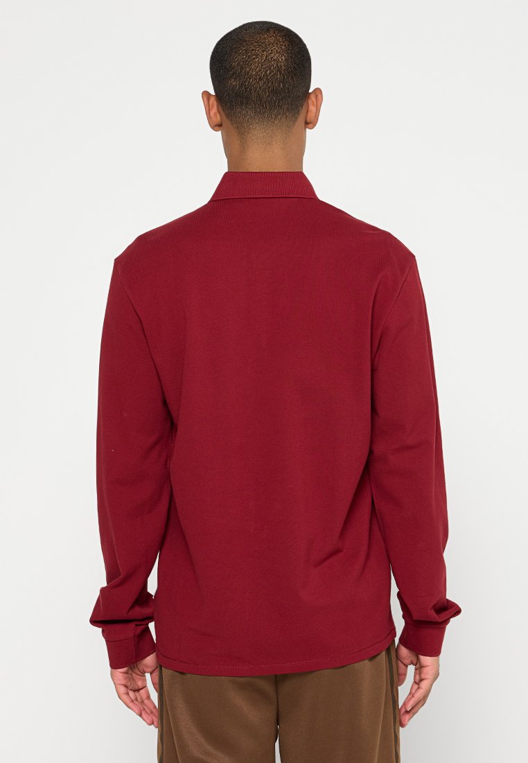 Młody mężczyzna widziany z tyłu, ubrany w bordową koszulę z długim rękawem i brązowe spodnie na jednolitym białym tle.