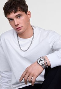 Giovane uomo in una camicia bianca a maniche lunghe, con una collana d'argento, un orologio d'argento e un bracciale d'argento abbinato, seduto su uno sfondo semplice.