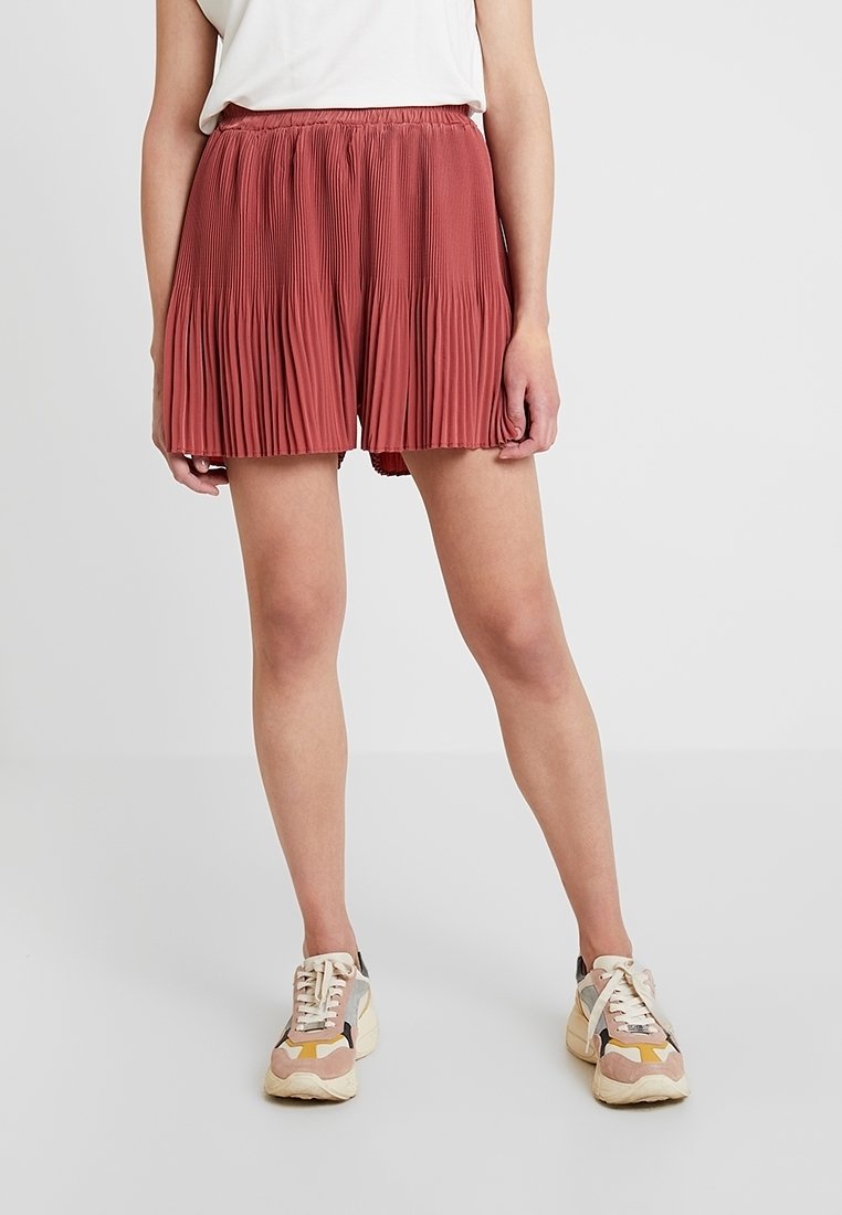 Shorts plissés de couleur rouille avec une taille élastique, fabriqués en tissu léger, associés à des baskets multicolores.