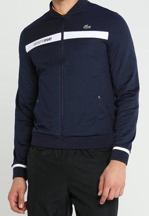 Giacca navy con zip e collo alto, caratterizzata da dettagli a righe bianche e nere e logo Lacoste. Realizzata in materiale liscio e leggero.
