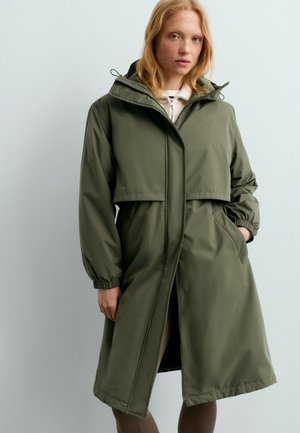 Jeune femme aux cheveux roux portant un long imperméable à capuche vert olive avec des poignets élastiques et les mains dans les poches sur un fond pâle.