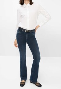 Femme portant une chemise blanche à boutons, un jean bootcut bleu foncé avec une ceinture noire, et des ballerines noires en résille, debout avec une main sur la hanche.