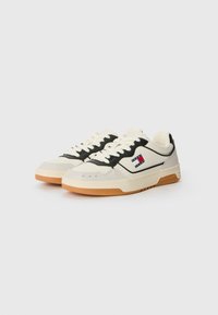 Cream and black sneakers with a textured finish, featuring lace-up closure, a logo patch on the side, and a gum sole.