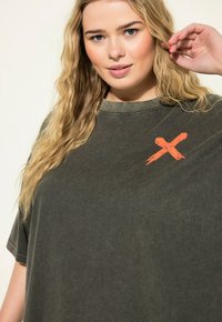 Olivengrünes, übergroßes T-Shirt mit rundem Halsausschnitt und einem auffälligen orangen Pinselstrich-'X' auf der Brust. Weicher, strukturierten Stoff.