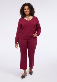 Maglione bordeaux a maglia con scollo a V, abbinato a pantaloni slim-fit bordeaux. Il modello indossa ballerine marroni, caratterizzate da un tessuto strutturato e una vestibilità rilassata.