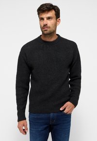 Schwarzer strukturierter Pullover mit einem Chevron-Muster, geripptem Ausschnitt und Säumen. Kombiniert mit blauen Jeans im Straight-Fit-Design.