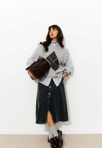 Gilet en maille gris avec un motif en losange sur une chemise rayée bleu clair, associé à une jupe en velours côtelé noir et des mocassins marron, portant un sac marron.
