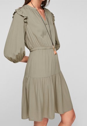 Kvinde iført en lys olivengrøn kjole med ballonsærmer, flæsedetaljer på skuldrene, en elastisk talje og en sort vedhængshalskæde.