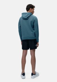 Hooded pulover v potlačeni teal barvi, v kombinaciji s črnimi kratkimi hlačami. Pulover ima mehak teksturo in rebraste manšete, z klasičnim, sproščenim oblikovanjem.