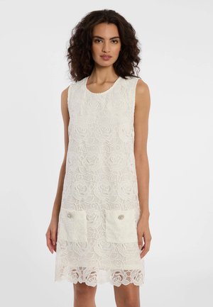 Frau mit lockigem Haar trägt ein ärmelloses weißes Spitzenkleid mit Blumenmustern und zwei vorderen Taschen, steht vor einem einfarbigen Hintergrund.