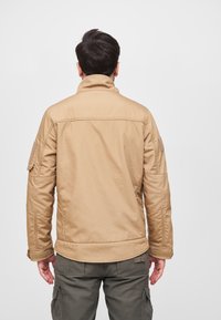 Khaki jacket with a high collar, textured fabric, side pockets, and military-style accents. Worn with olive cargo pants.