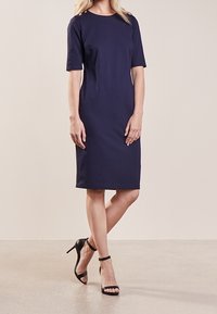 Žena v námořnicky modrých šatech ke kolenům s krátkými rukávy a černými sandály na vysokém podpatku, stojí s jednou nohou mírně překříženou.