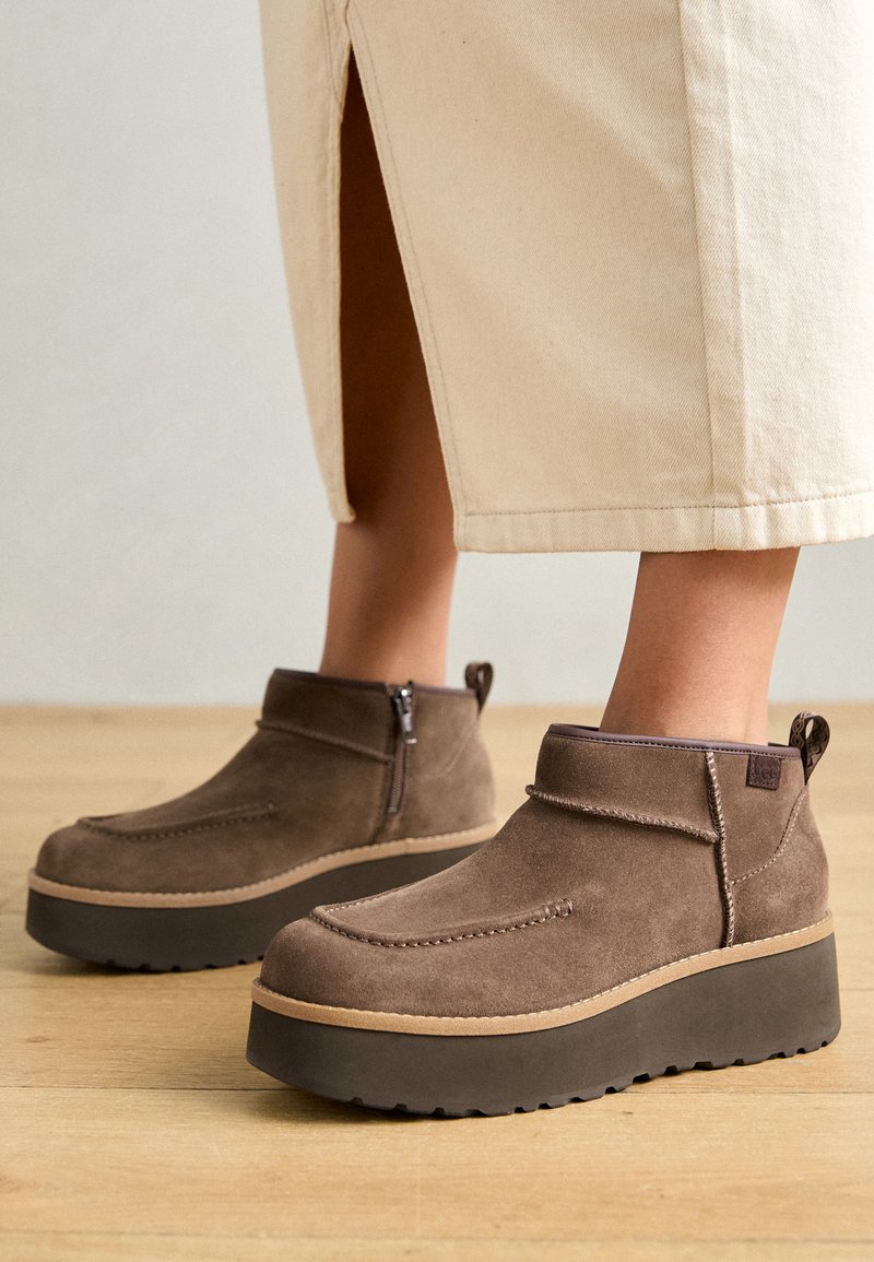 Braune Wildleder-Ankle-Boots mit einer klobigen dunklen Plattformsohle, seitlichem Reißverschluss und dekorativen Nähten. Auf einem hellen Holzboden präsentiert.
