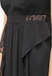 Robe noire avec taille en dentelle et panneaux en tissu transparent à volants sur les côtés, montrée de la taille jusqu'à la partie supérieure de la cuisse.