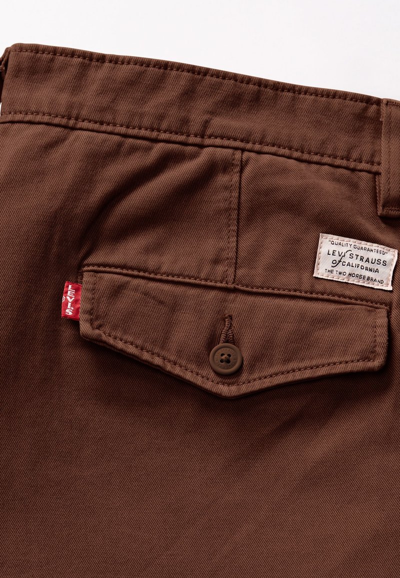 Tessuto di cotone twill marrone con una tasca posteriore caratterizzata da un bottone marrone e un'etichetta Levi's con testo nero e un accento rosso sulla cucitura laterale.