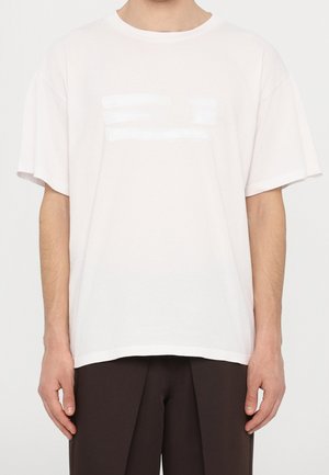 Mężczyzna ubrany w oversize'ową, gładką, białą koszulkę i ciemnobrązowe spodnie, stojący z ramionami luźno opuszczonymi wzdłuż ciała na białym tle.