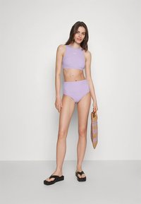 Lilla to-delt bikini med crop-top og høy midje på underdelen. Modellen holder en stripet vevd veske og har på seg svarte flip-flops.