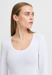White long-sleeve top with a rounded neckline. Smooth texture, fitted design, and minimalistic style. Model has light brown hair and small earrings.