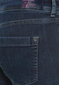 Mörkblå denimjeans med en bakficka med en metallnitar, robust twilltyg och kontrasterande gula sömmar.