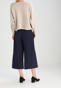 Mujer vistiendo un suéter de manga larga beige, pantalones cortos de pierna ancha en color azul marino y zapatos de tacón cuadrado negros, de pie frente a un fondo liso.