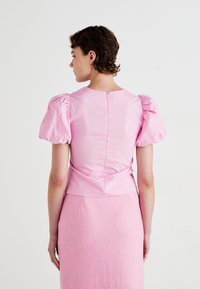 Kort rosa bluse med puffede ærmer, glat tekstur og en lynlås i ryggen, parret med en matchende struktureret rosa nederdel.