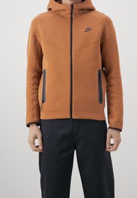 Veste à capuche Nike en orange, dotée d'une fermeture éclair à l'avant, de deux poches latérales avec zippers, et de accents noirs aux poignets et aux bordures des poches.
