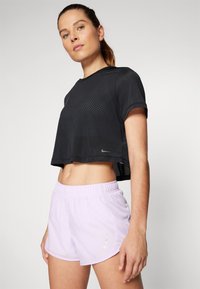 Melnā cropped mežģu sporta augšdaļa ar īsām piedurknēm, kopā ar gaiši purpursarkanām šortiem ar elastīgu jostasvietu un atstarojošu logo detaļu.