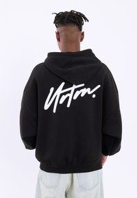 Felpa nera con vestibilità over size, caratterizzata da un logo script bianco sul retro. Realizzata in tessuto morbido, include un cappuccio con stringhe e orlo a coste.