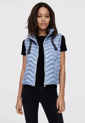 Donna che indossa un gilet trapuntato azzurro chiaro senza maniche sopra una maglietta nera a maniche corte e pantaloni neri, in piedi su uno sfondo semplice.