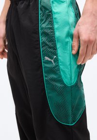 Κοντινό πλάνο μαύρου και τιρκουάζ παντελονιού φόρμας Puma με σχεδιασμένη τιρκουάζ πλαϊνή λωρίδα και λογότυπο Puma κοντά στην τσέπη, φορεμένου από άτομο.