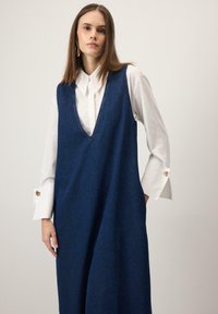 Tumši zils džinsa kombinezons ar V veida izgriezumu. Zem tā valkāta balta garām piedurknēm krekls ar pogājamiem manšetiem. Gluda tekstūra, brīvs siluets.