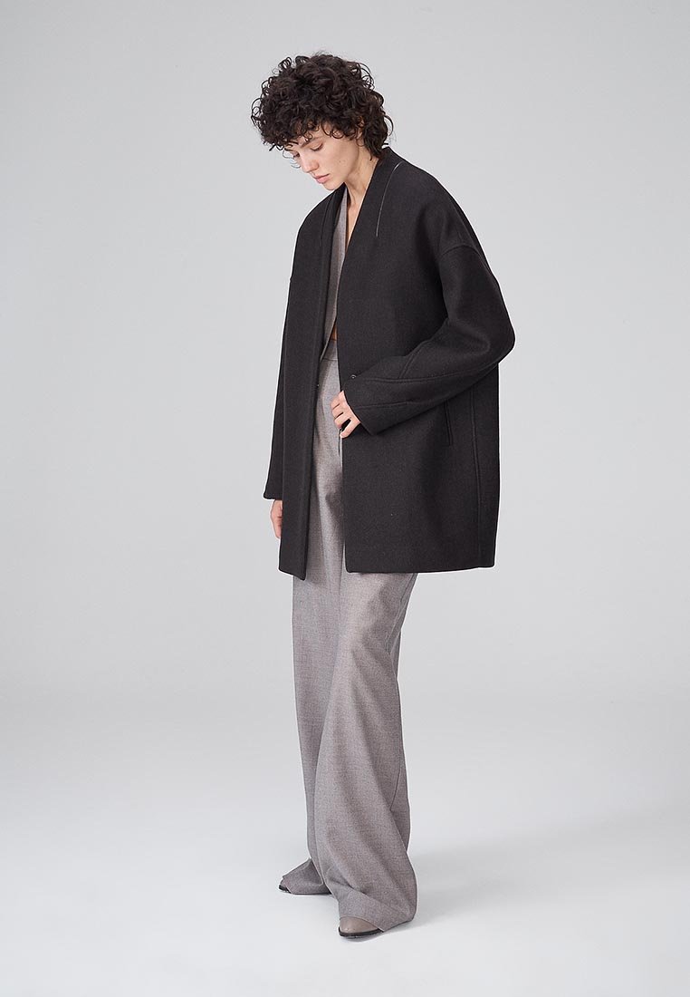 Persona con capelli ricci che indossa un cappotto nero oversize sopra pantaloni grigi a gamba larga, in piedi e che guarda in basso contro uno sfondo grigio semplice.