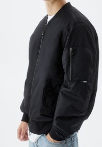 Veste bomber noire en tissu lisse, avec une fermeture éclair à l'avant, des poches latérales, et une poche zippée distincte sur la manche gauche.