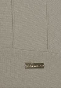 Detailní záběr béžové tkaniny s obdélníkovou kovovou nápisovou štítkou s raženým značkovým názvem "LASCANA" přišitým k materiálu.