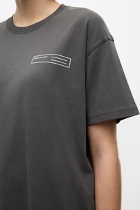 Dark gray short-sleeve t-shirt made of cotton, featuring a white printed label design with lines on the upper left. Soft texture, loose fit.