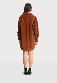 Braunes Cordhemdkleid mit Kragen, langen Ärmeln und einem rückseitigen Faltendetail. Glatte Textur und knielang mit geknöpften Manschetten.